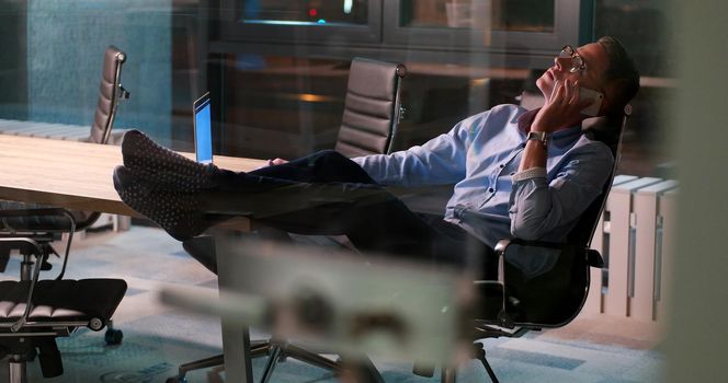 Full length of a relaxed casual young businessman sitting with legs on desk at night office