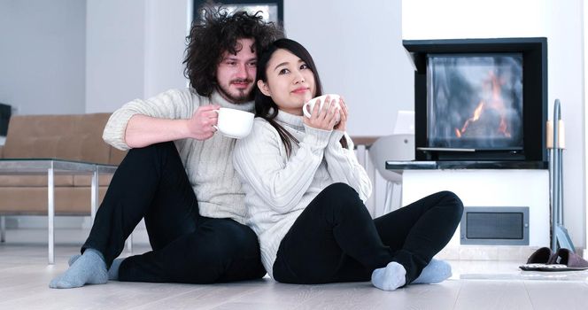 Young romantic multiethnic couple sitting on the floor in front of fireplace at home, looking at each other, talking and drinking coffee at autumn day