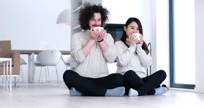 Young romantic multiethnic couple sitting on the floor in front of fireplace at home, looking at each other, talking and drinking coffee at autumn day