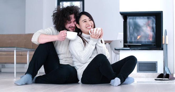 Young romantic multiethnic couple sitting on the floor in front of fireplace at home, looking at each other, talking and drinking coffee at autumn day