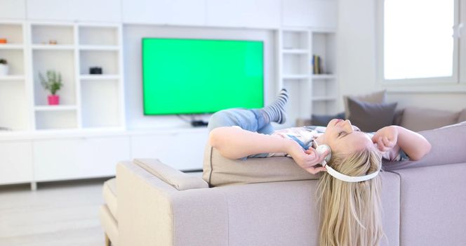 Young beautiful girl enjoying music through headphones, laying on sofa at home