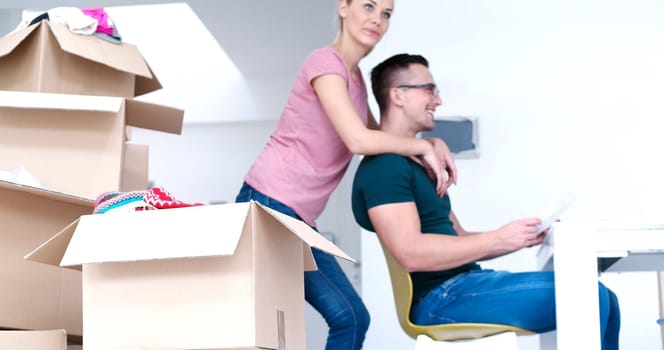 Young couple moving in a new home. Man and woman at the table using notebook laptop computer and plans with boxes around them