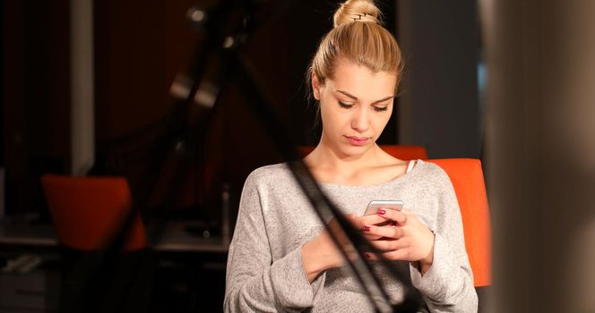 Young woman using mobile phone while working on computer at night in dark office.