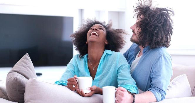 young multiethnic couple sitting on sofa at home drinking coffe, talking, smiling.