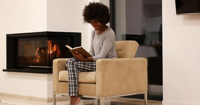 Young beautiful african american woman sitting in front of fireplace at home on a cold autumn day and reading book