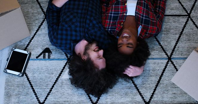 Top view of attractive young multiethnic couple moving in, holding hands, looking at camera and smiling while lying among cardboard boxes