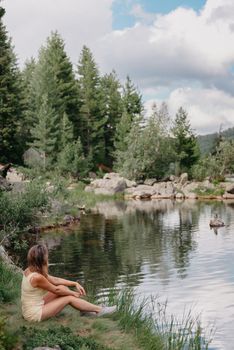 Tourist girl enjoys the magical view of the lake, coniferous forest and magical view sitting on big stone on the shore of a turquoise lake in the mountains. Hiking in the Natural Park. Black lake.