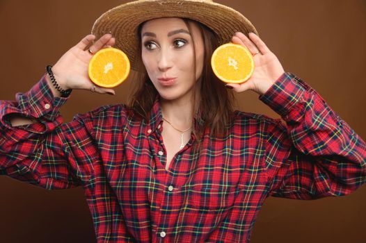 Attractive rustic Caucasian young woman in straw hat holding cut orange fruit. Studio portrait over brown background. Looks into the camera.