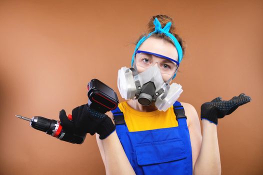 Portrait of a caucasian young woman in the uniform of a superintendent of streetwear in overalls, respirator and goggles with a screwdriver in hand. Looks into the camera. In annoyance, he throws up his hands after a mistake or misunderstanding. Builder bug concept