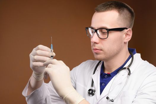 Young caucasian doctor man preparing to do a vaccination with a syringe. Studio shot on a brown background. Release air from the syringe before the injection.