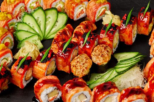 Closeup of set of delicious rolls, nigiri sushi and gunkan maki with salmon garnished with tobiko, fresh cucumber, shredded daikon and pickled ginger on black background. Japanese cuisine