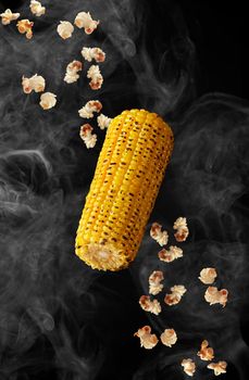 Grilled ear of corn and popped corn kernels hovering in white smoke isolated on black background. Popular organic snacks concept