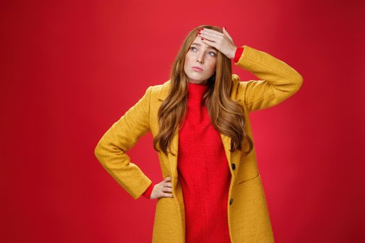 Girl having feaver touching forehead with palm looking displeased and bothered left as thinking she got cold standing in yellow coat, realising she sick or ill over red background, feeling tired.