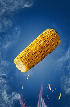 Yellow grilled ear of corn hovering over flames in sparks and white haze on blue background. Natural food concept. Vertical image with copyspace