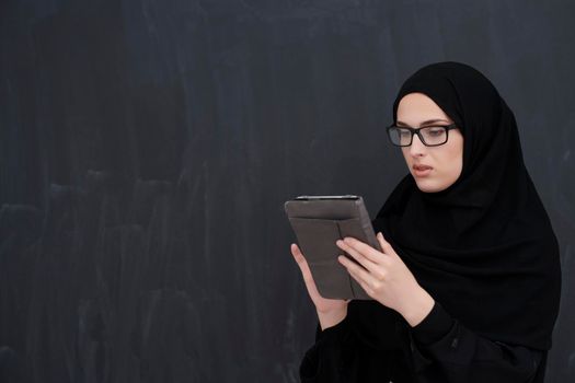 Young Arab businesswoman in traditional clothes or abaya and glasses holding tablet computer in front of black chalkboard representing modern islam fashion and technology