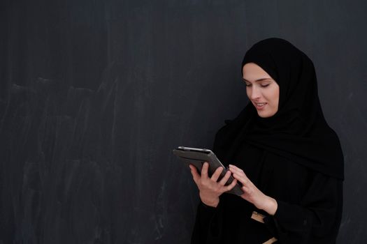 Young Arab businesswoman in traditional clothes or abaya and glasses holding tablet computer in front of black chalkboard representing modern islam fashion and technology