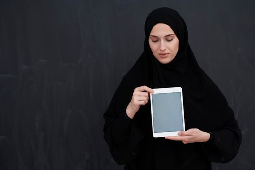 Young Arab businesswoman in traditional clothes or abaya and glasses holding tablet computer in front of black chalkboard representing modern islam fashion and technology