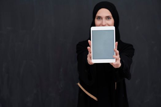 Young Arab businesswoman in traditional clothes or abaya and glasses holding tablet computer in front of black chalkboard representing modern islam fashion and technology