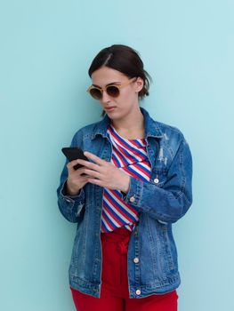 Portrait of young girl using smartphone while standing in front of blue background. Female model wearing sunglasses representing modern fashion and technology concept