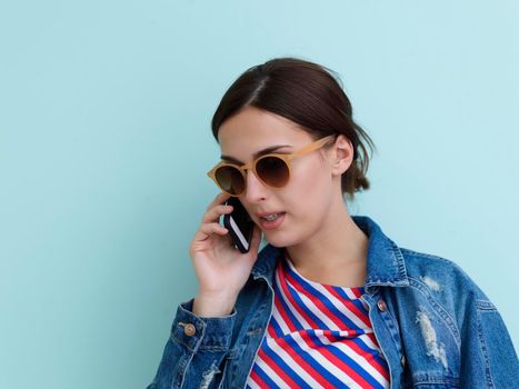 Portrait of young girl talking on the phone while standing in front of blue background. Female model wearing sunglasses representing modern fashion and technology concept