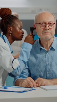 Young specialist doing ear consultation with otoscope on sick old patient in examination office. Otologist using medical instrument for checkup and professional analysis on elder man