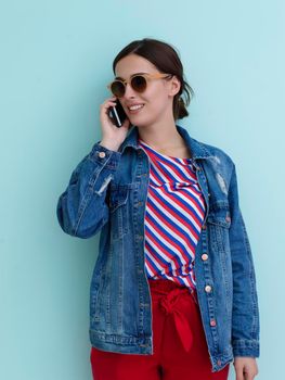 Portrait of young girl talking on the phone while standing in front of blue background. Female model wearing sunglasses representing modern fashion and technology concept