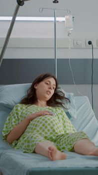 Pregnant woman laying in hospital ward bed waiting on assistance for childbirth delivery. Caucasian man holding backpack for wife expecting child sitting together at clinic to give birth