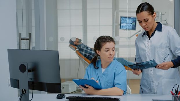 Professional team of dentist and nurse working on patient teethcare using dental x ray scan, stomatology equipment and computer at dentistry clinic. Orthodontists with medical tools