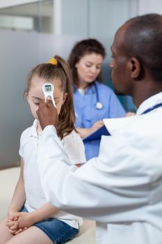 African american therapist pediatrician doctor measuring temperature of little child using medical infrared thermometer during clinical examination. Practitioner working in hospital office