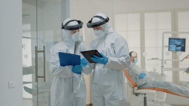 Dentists in protection suits talking about medicine before starting consultation with patient in dental cabinet. Caucasian team of dentistry specialists discussing dental surgery operation