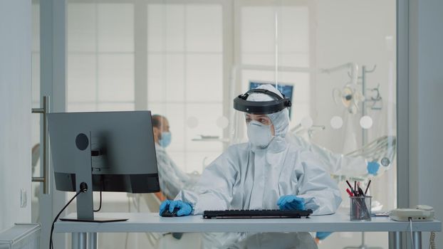 Portrait of dental assistent typing on computer keyboard wearing protection uniform with face shield, mask, gloves and coverall. Stomatology nurse using monitor technology at oral clinic