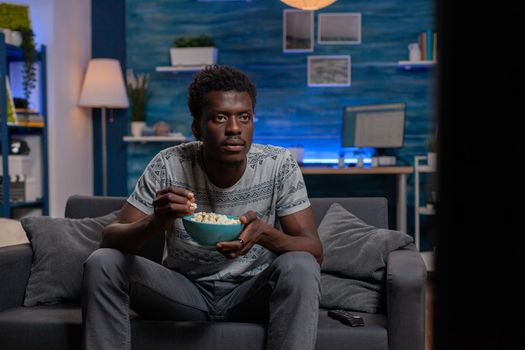 African american young man looking at football game on television sitting on couch on living room Guy watching sport channel on TV enjoying popcorn snack during free time activity at home