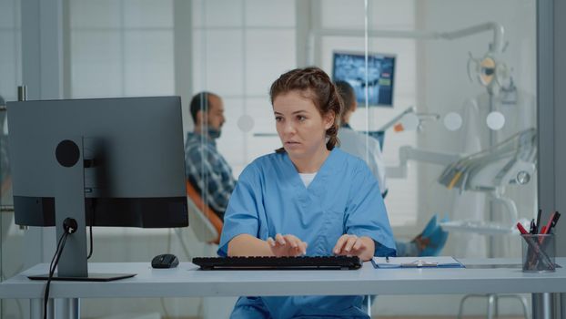 Dental assistant using computer and x ray scans at desk while stomatologist examining patient teethcare. Caucasian staff of orthodontists working at oral clinic for stomatology care