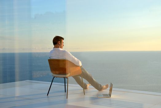 handsome young man relaxing and working on laptop computer at home balcony while looking sunset