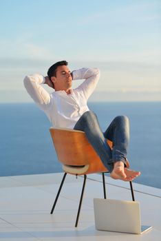 handsome young man relaxing and working on laptop computer at home balcony while looking sunset