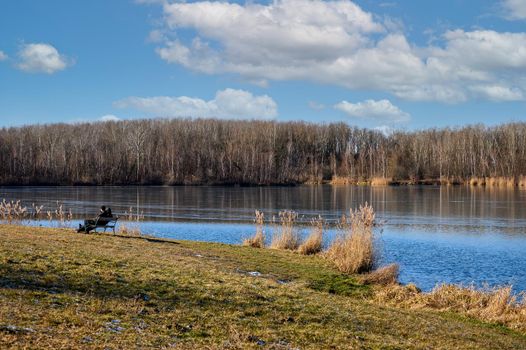 A couple is sitting on a bench and enjoying the first rays of spring sunshine on a river bank.
