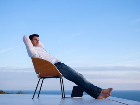 handsome young man relaxing and working on laptop computer at home balcony while looking sunset