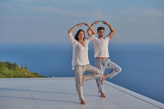 young couple practicing yoga at sunset in modern home terace with ocean and sunset in background