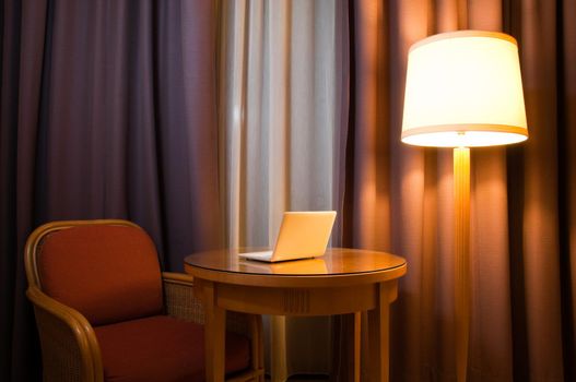 notebook computer on the table in the room with lamp, chair and curtains on the window