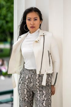 A young pretty Mongolian woman with a white leather jacket and patterned well-fitting pants in an urban park.