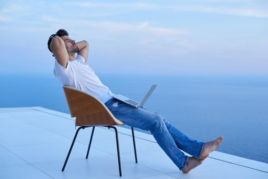 handsome young man relaxing and working on laptop computer at home balcony while looking sunset