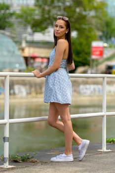 A portrait of a pretty young woman, taken in the summer in a city, leaning against a railing on a nearby river.