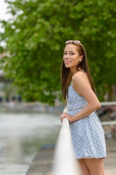 A portrait of a pretty young woman, taken in the summer in a city, leaning against a railing on a nearby river.
