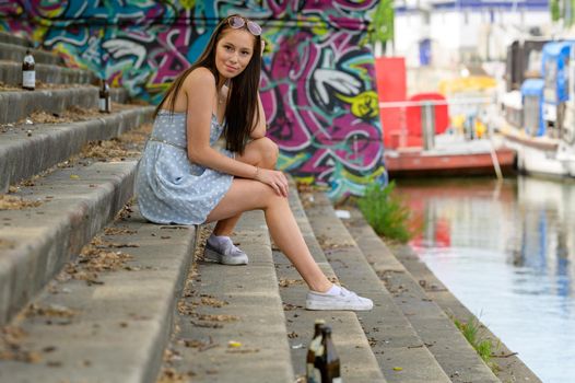 A portrait of a pretty young woman taken in the summertime in a city sitting on a staircase next to a river.