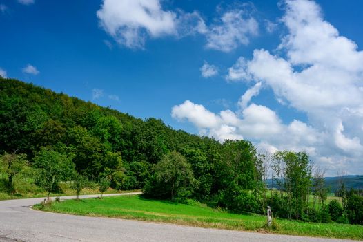 A narrow country road blends into the lush green summer landscape and is framed by a deep blue sky with a few white clouds.