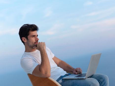 handsome young man relaxing and working on laptop computer at home balcony while looking sunset