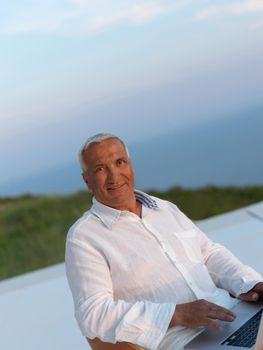 relaxed white hair senior man looking sunset from luxury balclony home and working on laptop computer