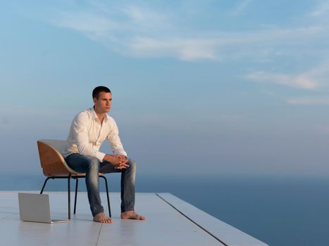 handsome young man relaxing and working on laptop computer at home balcony while looking sunset