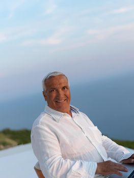 relaxed white hair senior man looking sunset from luxury balclony home and working on laptop computer