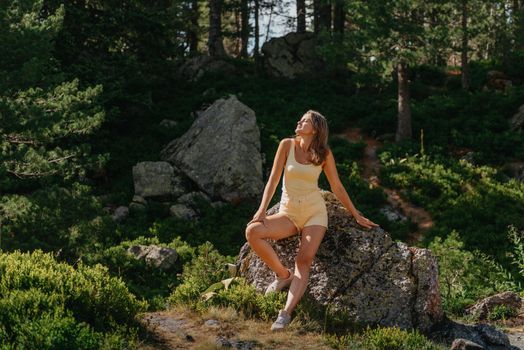 Pretty young girl in the yellow T-shirt and yellow shorts with sunglasses sitting on the stone above the beautiful canyon with an emerald lake on the sunset in Spain. Traveler in the mountains.